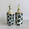 Two ceramic oil cruets with gold pour spouts. The cruets feature off-white and black floral designs in varying sizes.