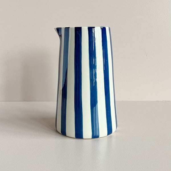 A ceramic handle-less pitcher jug in bold navy blue and natural white stripes against a neutral backdrop.