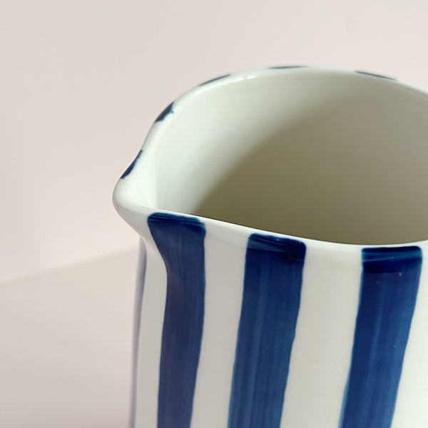 A close up view of the pour spout of a ceramic blue and natural white striped jug, against a neutral backdrop.