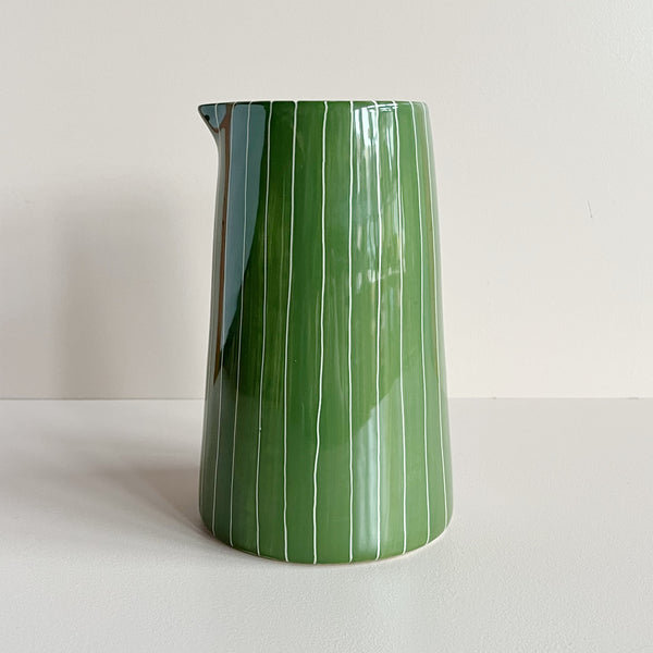 A ceramic handleless pitcher jug featuring a vibrant moss green and narrow white striped pattern, sitting against a neutral backdrop.