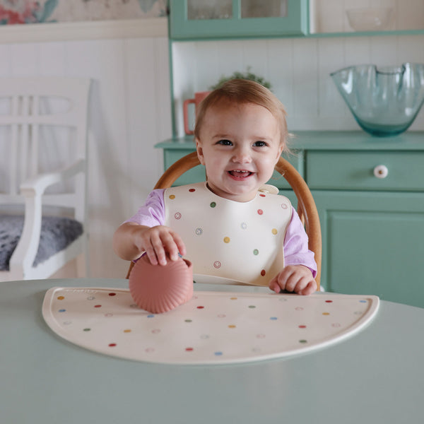 Child in a high chair with a pink cup and colorful placemat in a kitchen setting