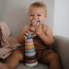 Baby sitting on a couch holding a colorful stacking toy and a pacifier.