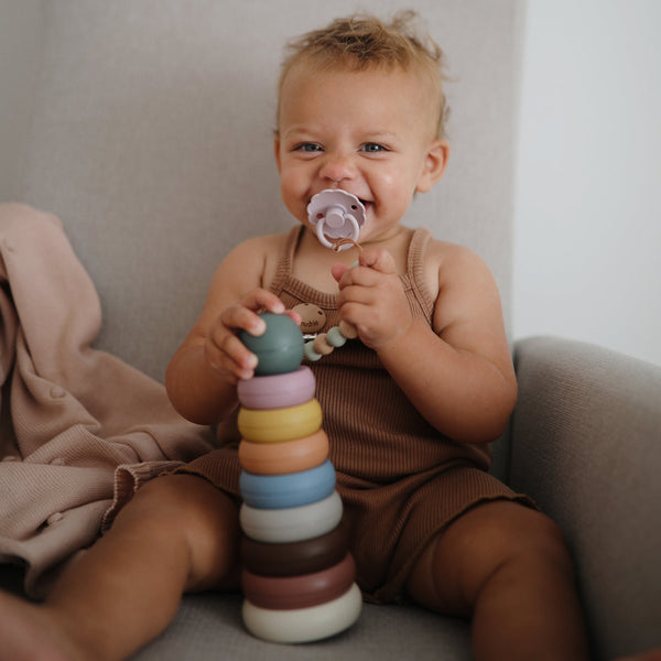 Baby sitting on a couch holding a colorful stacking toy and a pacifier.