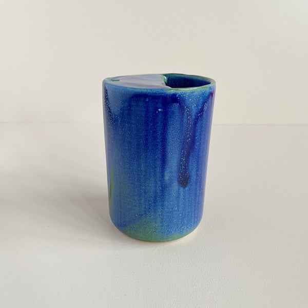 A small ceramic to-go cup featuring a vibrant blue glaze with subtle pops of green photographed against a neutral backdrop.