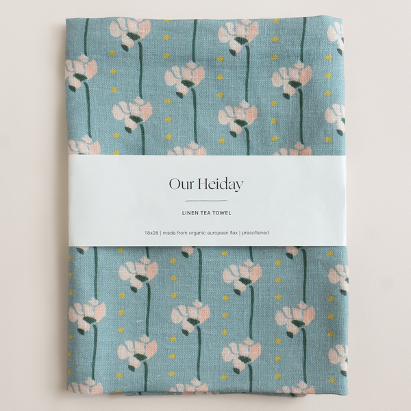 Linen tea towel with blue floral pattern and 'Our Heiday' branding on a light beige background