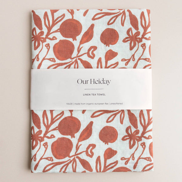 Tea towel with rust red pomegranate pattern on a white background, branded 'Our Heiday'.
