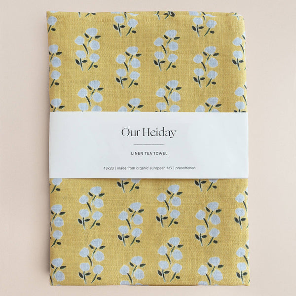 Yellow linen tea towel with floral pattern and 'Our Heiday' label on a beige background