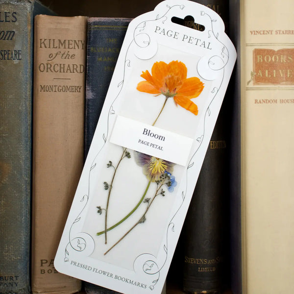 Packaging from 'Page Petal' containing a clear bookmark with a 'Bloom' theme featuring pressed orange and purple flowers, leaning against a backdrop of old books.