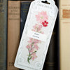 A decorative pressed flower bookmark featuring pressed pink and red flowers, leaning against a row of vintage books on a wooden shelf. The white packaging reads 'PAGE PETAL' and 'Love Page Petal'.