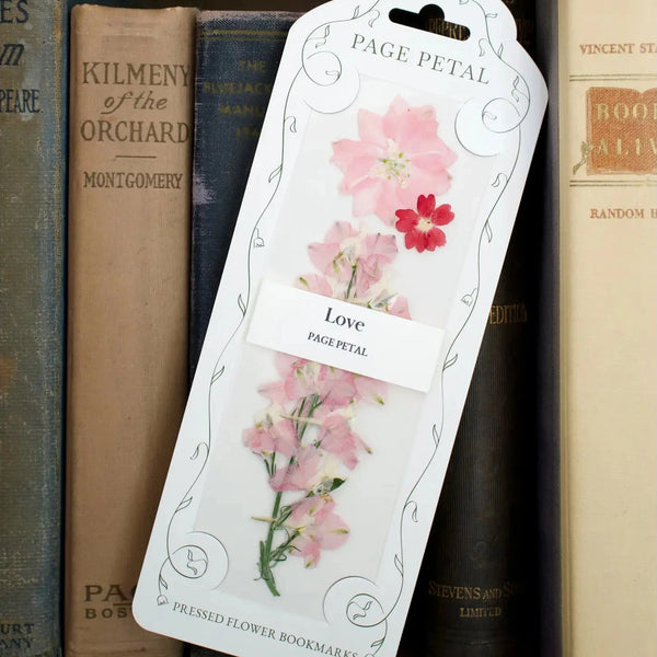 A decorative pressed flower bookmark featuring pressed pink and red flowers, leaning against a row of vintage books on a wooden shelf. The white packaging reads 'PAGE PETAL' and 'Love Page Petal'.