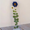 A clear bookmark containing a pressed green eucalyptus stem with a blue daisy and a small white daisy at the top, displayed on a wooden surface against a plain beige wall.