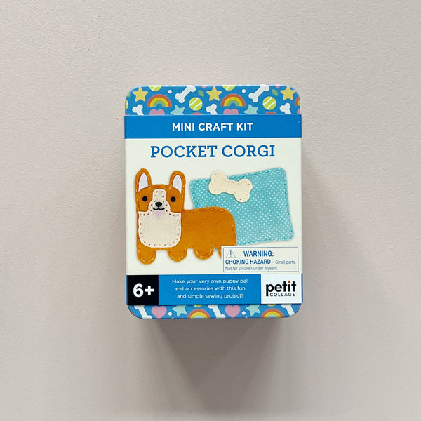 A Petit Collage 'Pocket Corgi' mini craft kit in its original packaging, shown against a neutral light gray surface. The box features a colorful design with a blue top and bottom border, a white center, and an illustration of a corgi and a blue fabric pillow.
