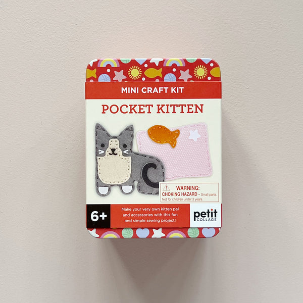 A Petit Collage 'Mini Craft Kit Pocket Kitten' in a square metal tin, mounted on a light beige wall. The packaging features a white center with illustrations of a gray and cream felt kitten and a pink felt fish, surrounded by a red top and bottom band with colorful patterned dots.