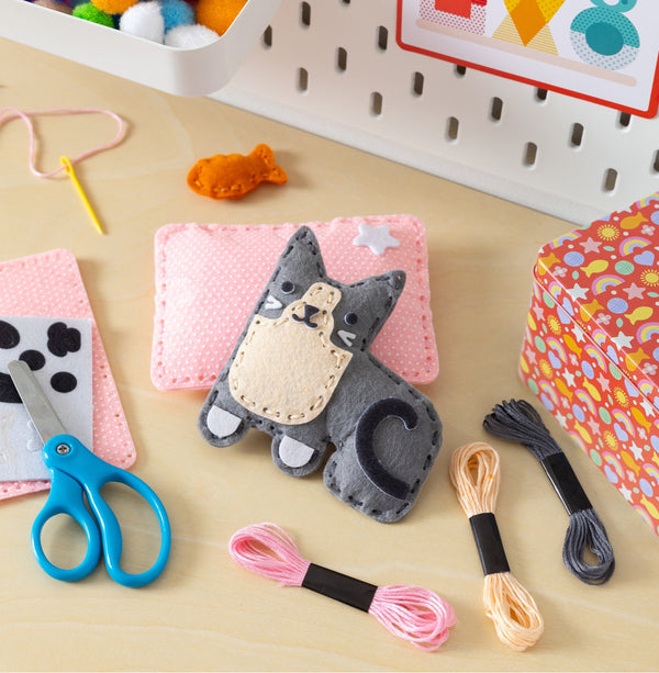 A felt kitten craft project laid out with embroiderly floss, safety scissors, and a kid-safe needle on a pale yellow craft table.