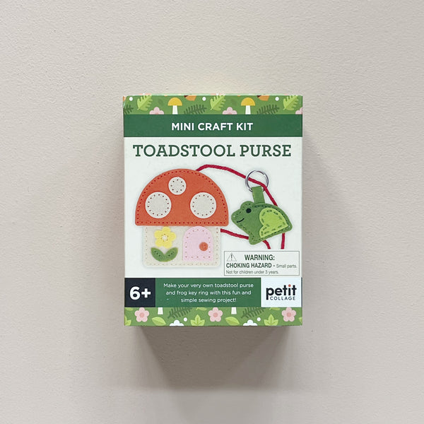A boxed mini craft kit labeled 'TOADSTOOL PURSE' by Petit Collage, shown against a plain light wall. The package features a mushroom-shaped purse and frog bag charm, with text including '6+' and a choking hazard warning.