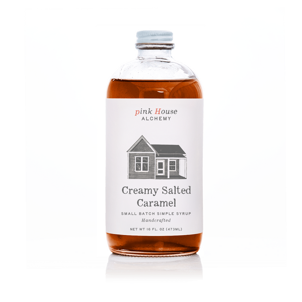 A bottle of Pink House Alchemy Creamy Salted Caramel syrup on a white background. The clear glass bottle shows the amber-brown syrup inside, with a white label featuring a simple house illustration and the brand name 'pink House ALCHEMY'.