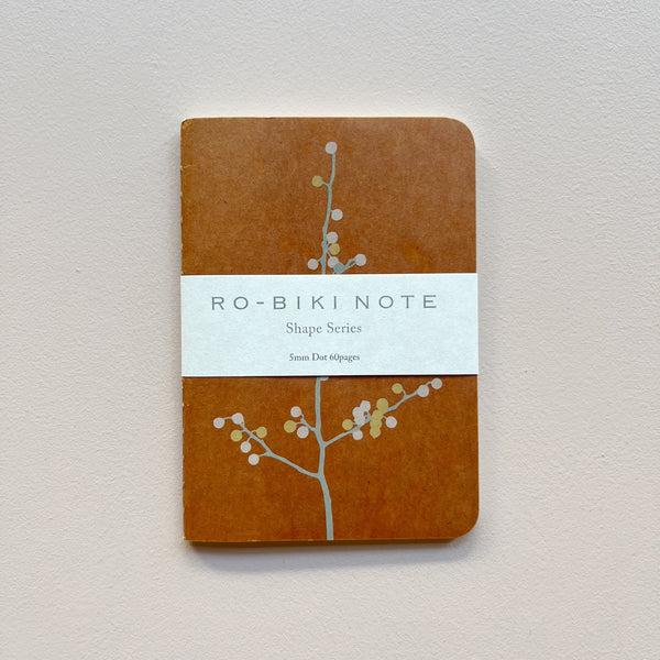 A small notebook featuring a warm caramel brown waxed paper cover featuring a design of pastel flowering tree with white packaging saying 'Ro-biki Note Shape Series' against a neutral background.