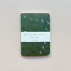 A small notebook featuring a dark green waxed paper cover featuring a design of pastel flying birds with white packaging saying 'Ro-biki Note Shape Series' against a neutral background.