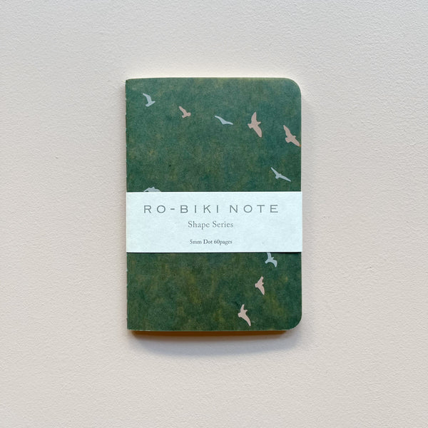 A small notebook featuring a dark green waxed paper cover featuring a design of pastel flying birds with white packaging saying 'Ro-biki Note Shape Series' against a neutral background.