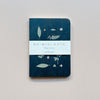 A small notebook featuring a dark blue waxed paper cover featuring a design of pastel wild plants with white packaging saying 'Ro-biki Note Shape Series' against a neutral background.