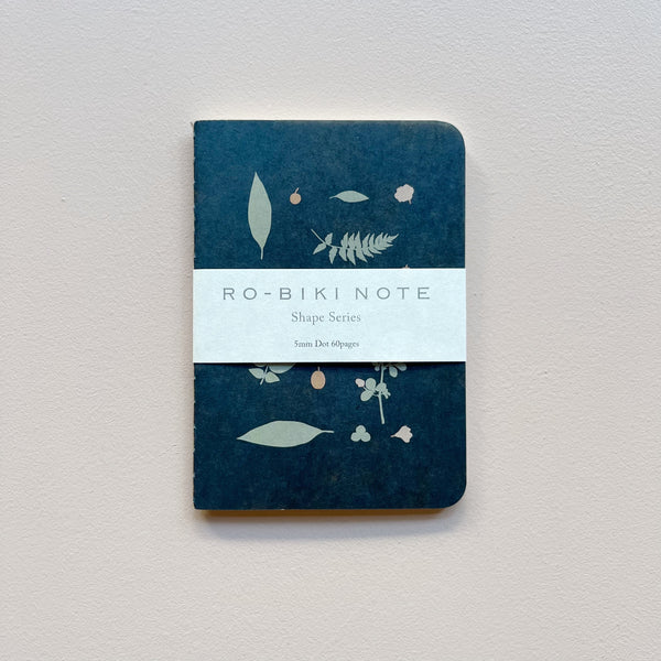 A small notebook featuring a dark blue waxed paper cover featuring a design of pastel wild plants with white packaging saying 'Ro-biki Note Shape Series' against a neutral background.