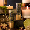 Two cylindrical lip balm tubes from Poppy & Pout stand on a dark wooden surface in a styled setting with lit candles, dried flowers, greenery, and a lime slice. The left tube is labeled Midnight Margarita and the right tube is labeled Garden Spell.