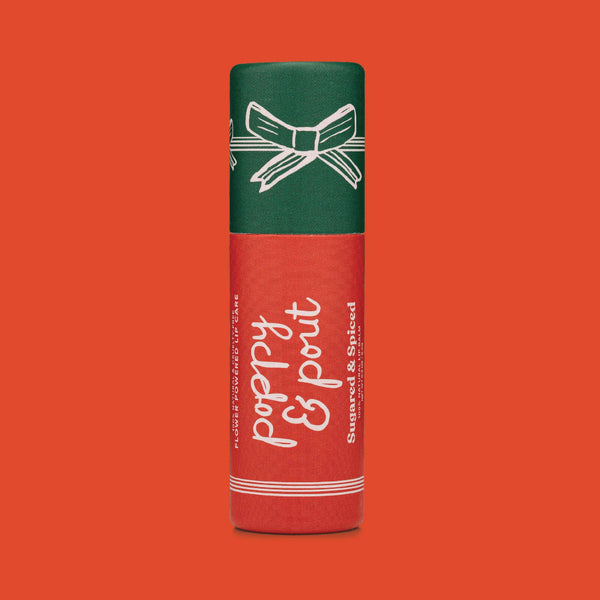 A single lip balm tube standing upright on a solid orange-red background. The tube is dark green on top with a white bow illustration and white text reading 'poppy & pout', with additional text 'Sugared & Spiced'.