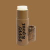A lip balm stick with its cap off, standing next to it on a solid brown background. The balm stick has a white top and a brown paper sleeve printed with 'poppy & pout' and 'Vanilla Chai'.