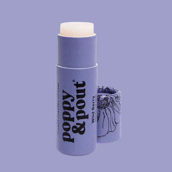 A lavender lip balm stick with its cap removed and standing beside it on a matching lavender background. The balm stick has a pale cream-colored balm tip and black text reading 'poppy & pout' and 'wild Berry'.