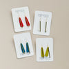 Four pairs of long teardrop clay and brass earrings on cards labeled 'Poppyseed' against a beige background
