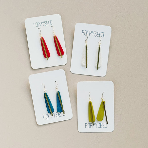 Four pairs of long teardrop clay and brass earrings on cards labeled 'Poppyseed' against a beige background