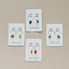 A display of four different colors of gold hoop earrings with a seed shaped clay drop alongside a small brass accent.