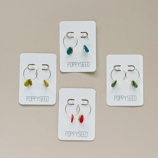 A display of four different colors of gold hoop earrings with a seed shaped clay drop alongside a small brass accent.