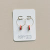Pair of hoop earrings with red clay and brass accents on a 'Poppyseed' branded card against a beige background