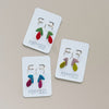 Three pairs of gold hoop and clay floral earrings on cards with 'Poppyseed' branding on a beige background