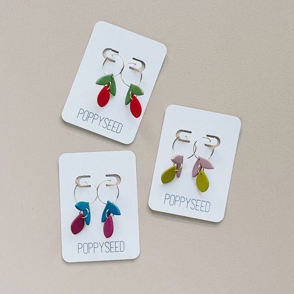 Three pairs of gold hoop and clay floral earrings on cards with 'Poppyseed' branding on a beige background