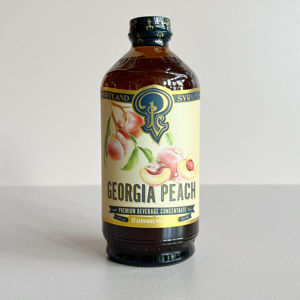 A bottle of Portland Georgia Peach Premium Beverage Concentrate syrup, featuring an amber glass bottle with a cream-colored label decorated with peach illustrations.