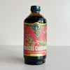 A bottle of Portland Syrup Hibiscus-Cardamom Premium Beverage Concentrate on a light gray background. The bottle is dark amber glass with a vibrant pink label featuring green leaves and red hibiscus flowers, and a blue cap.