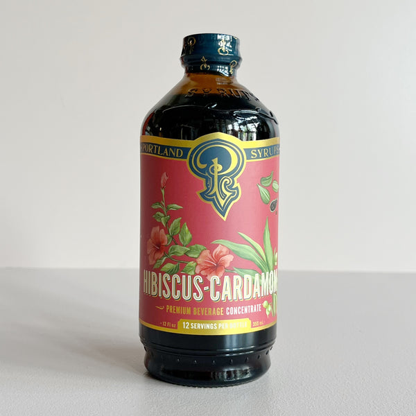 A bottle of Portland Syrup Hibiscus-Cardamom Premium Beverage Concentrate on a light gray background. The bottle is dark amber glass with a vibrant pink label featuring green leaves and red hibiscus flowers, and a blue cap.