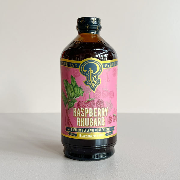 A bottle of Portland Syrup Raspberry Rhubarb Premium Beverage Concentrate, featuring a pink label with green and gold details, set against a plain light gray background.