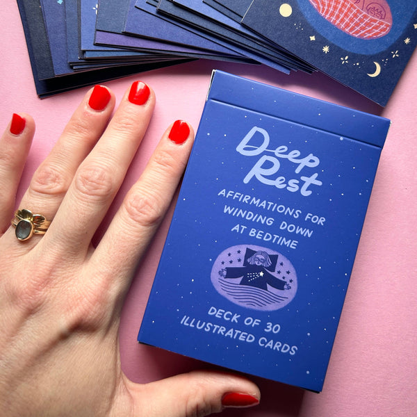 Blue card deck titled 'Deep Rest' held by a hand with red nail polish on a pink background