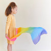 A child wearing a yellow dress playing with a rainbow Playsilk against a neutral backdrop.