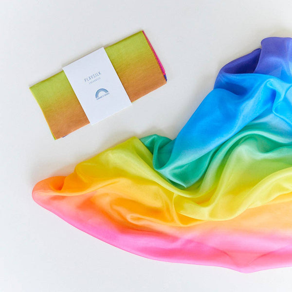 A rainbow-colored silk fabric square shown spread out against a white backdrop with another Playsilk folded into its packaging.