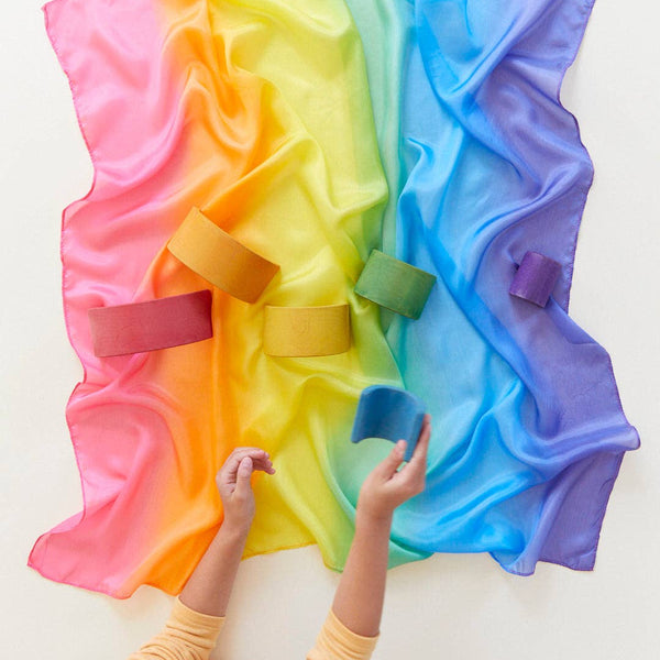A rainbow colored silk play square with block toys and child's hands playing, against a white backdrop.