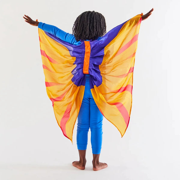 A child with locs in a blue outfit faces away with their arms spread showing off their silk pterodactyl wings.