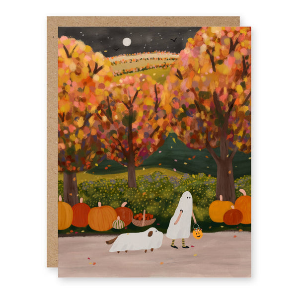 A greeting card with a kraft envelope behind it, featuring an autumn landscape illustration with colorful fall trees, pumpkins, a ghost figure, and a white dog on a light gray background.