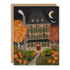 A Halloween themed greeting card depicting a spooky house decorated with jack'o'lanterns, garlands, and black cats, along with a skeleton, bat, ghost, and crescent moon. The card is paired with a kraft paper envelope.