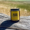 A small amber glass jar candle with a black lid and a bright yellow label reading 'BASE CAMP' sits on a weathered wooden surface outdoors, with a blurred field and blue sky in the background.