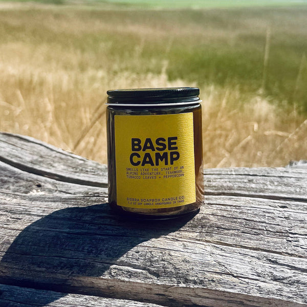 A small amber glass jar candle with a black lid and a bright yellow label reading 'BASE CAMP' sits on a weathered wooden surface outdoors, with a blurred field and blue sky in the background.
