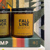 A brown glass jar candle with a golden yellow label with 'FALL LINE' and additional brand info listed below. It sits next to another candle atop a hardcover book and a blanket-covered wall.